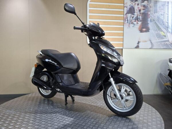 A black scooter with a sleek design is displayed on a circular metallic platform, set against a neutral background with a blurred image of people walking.