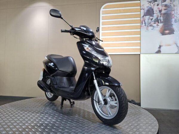 A black scooter is positioned on a circular silver platform against a neutral wall, with a contemporary backdrop featuring blurred pedestrians.
