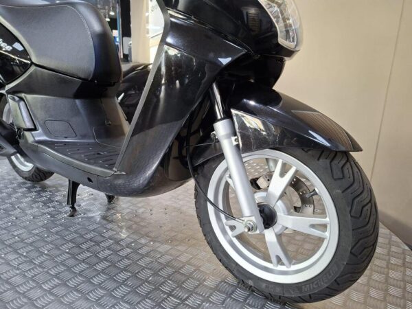 A close-up view of a black scooter, highlighting its front wheel, fork, and part of the seat, set against a textured flooring background.