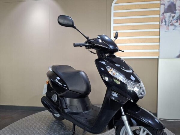 A black scooter positioned on a circular metal platform with a simple background, featuring light-colored walls and a partially visible graphic on the right.