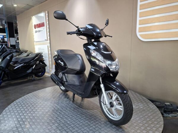 A black scooter is positioned on a circular display platform in a showroom, with other scooters visible in the background and a Yamaha sign on the wall.