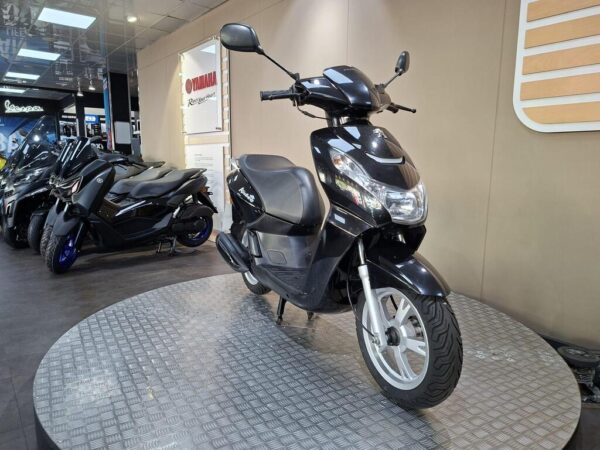 A black scooter displayed on a circular stand in a motorcycle showroom, with additional scooters in the background and a Yamaha sign visible.