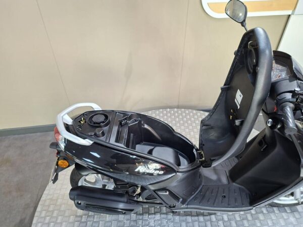 A black scooter with a partially open seat, revealing the storage compartment and fuel cap, positioned on a textured floor with a plain wall in the background.