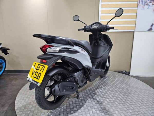 A silver scooter with a distinctive design parked on a textured circular platform, showcasing its rear and side profile. The license plate is visible, and the background features a neutral wall.