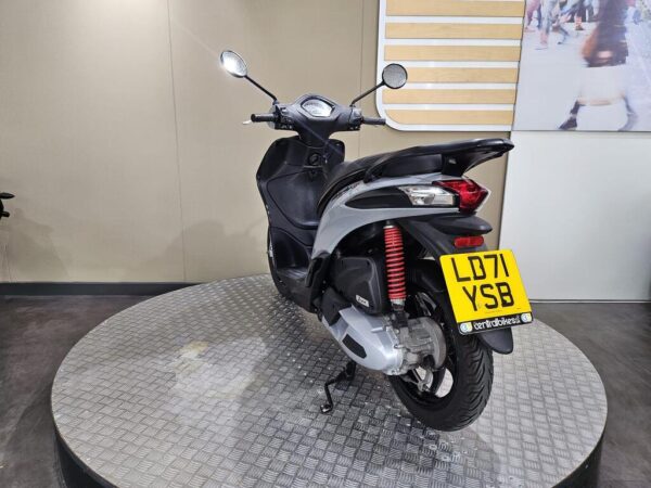 A black and silver scooter displayed on a round, textured platform, viewed from the rear. The scooter features a red shock absorber and has a license plate reading "LD71 YSB." The background includes a neutral wall with wood paneling.