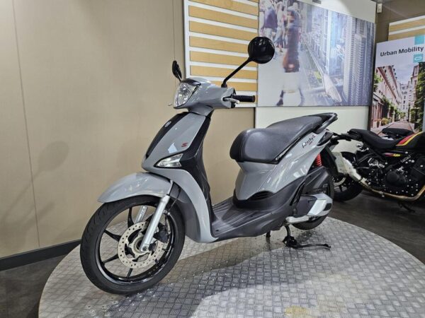 A gray scooter is displayed on a circular platform inside a showroom, with a modern background featuring urban imagery. Another motorcycle is partially visible in the background.
