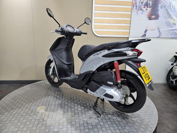 A silver and black scooter, model Vespa, is positioned on a textured circular platform with a simple background. The scooter is viewed from the rear-left angle, showcasing its sleek design and red shock absorbers.