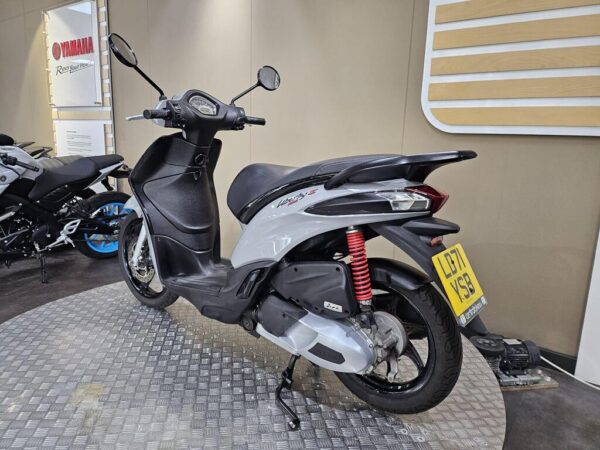 A Yamaha Neo's scooter displayed in a showroom, featuring a sleek gray body, red suspension spring, and a unique license plate; other motorcycles are visible in the background on a checkered floor.