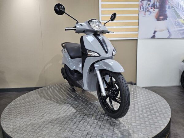 A light gray scooter is positioned on a textured circular platform, showcasing its sleek design and black wheels. The background features a simple, neutral wall.