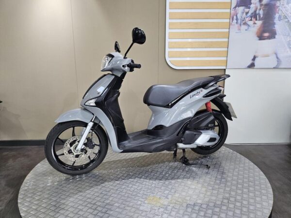 A grey scooter is displayed on a circular metal platform, with a neutral backdrop and a partially visible image of people walking in the background.