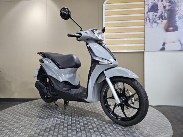 A gray scooter displayed on a circular metallic platform, featuring a sleek design, black wheels, and a modern look, with a blurred background of people walking.