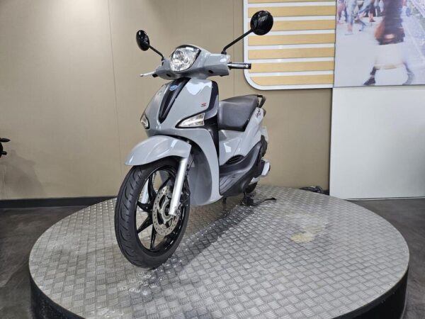 A gray scooter is displayed on a circular metal platform in a showroom setting, featuring a sleek design and modern aesthetics.