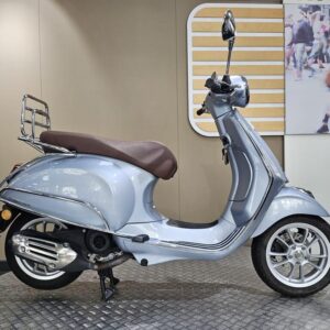 Vespa Primavera 50 (2023) A light blue scooter with a brown seat and chrome details, displayed in a sleek indoor setting with a patterned floor and a blurred background featuring a street scene.