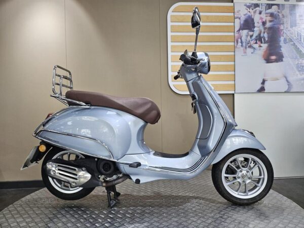 Vespa Primavera 50 (2023) A light blue scooter with a brown seat and chrome details, displayed in a sleek indoor setting with a patterned floor and a blurred background featuring a street scene.