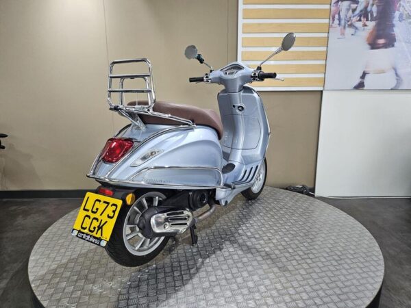 A silver scooter with a brown seat is displayed on a circular platform, featuring a rear luggage rack and visible license plate "LG73 CGK." The backdrop consists of neutral-colored walls and a large image of pedestrians.