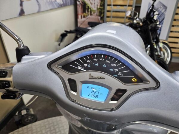 A close-up view of a scooter's speedometer and dashboard, featuring a speedometer gauge indicating kilometers per hour, a digital display showing total distance traveled, and warning lights on a silver panel. In the background, a blurred motorcycle is visible along with a decorative wall.