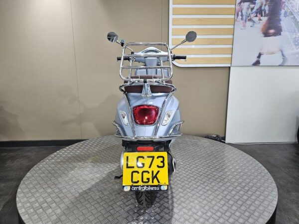 A rear view of a silver scooter displayed on a circular platform, featuring a chrome luggage rack, a red rear light, and a license plate with the registration "LC73 CGK." The background includes a minimalist wall with wooden panels and a partial view of a graphic.