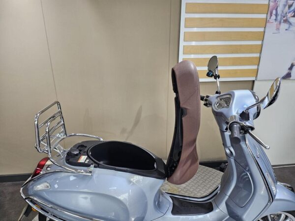 A silver scooter with a brown seat cover is parked against a plain wall, showcasing its chrome details and open storage area.