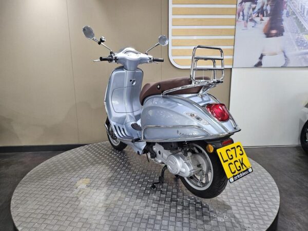 A silver Vespa scooter is displayed on a circular metallic platform, featuring a brown seat and a rear luggage rack. The background is a neutral-toned wall with a partial image of people walking.