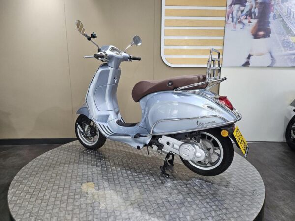 A light blue Vespa scooter with a brown seat is displayed on a circular metallic platform against a neutral wall. The scooter features chrome accents and a rear luggage rack.