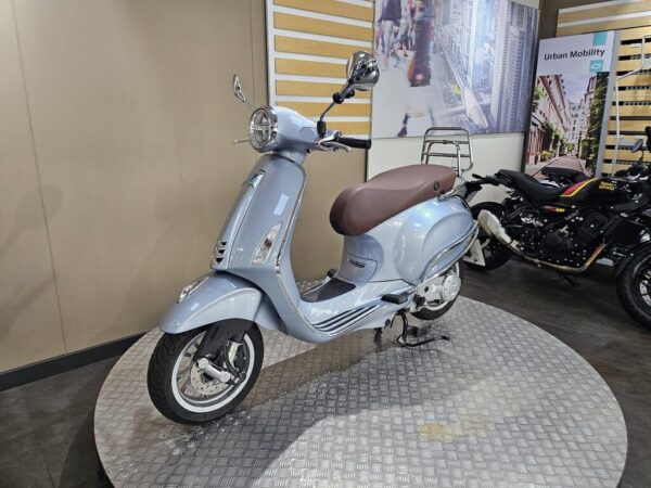 A light blue scooter with a brown seat is displayed on a circular platform, with a blurred background featuring other motorcycles and urban imagery on the walls.
