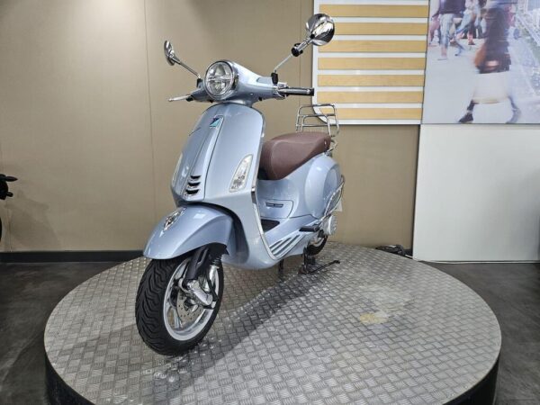 A light blue scooter with a brown seat, displayed on a circular metal platform, against a neutral background.