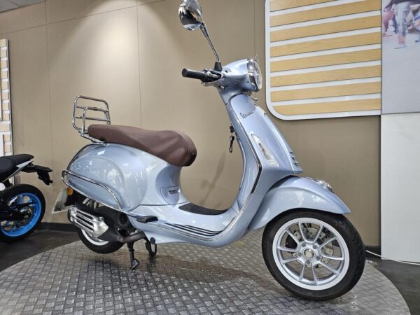 A light blue scooter with a brown seat and chrome details displayed in a showroom, featuring a rear luggage rack and stylish wheels.