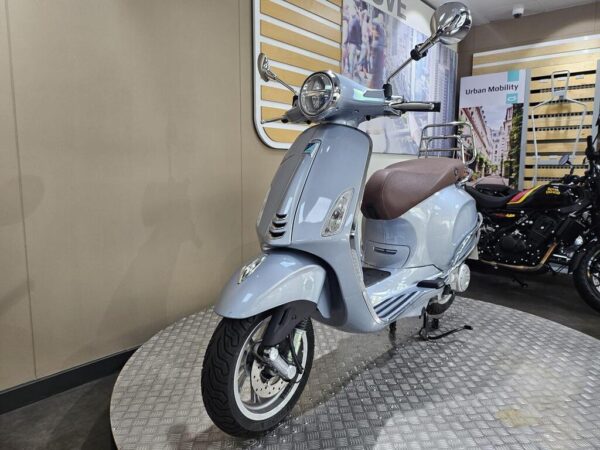A light blue vintage-style scooter with a brown seat displayed on a circular metallic platform, with a modern urban backdrop featuring promotional signage.