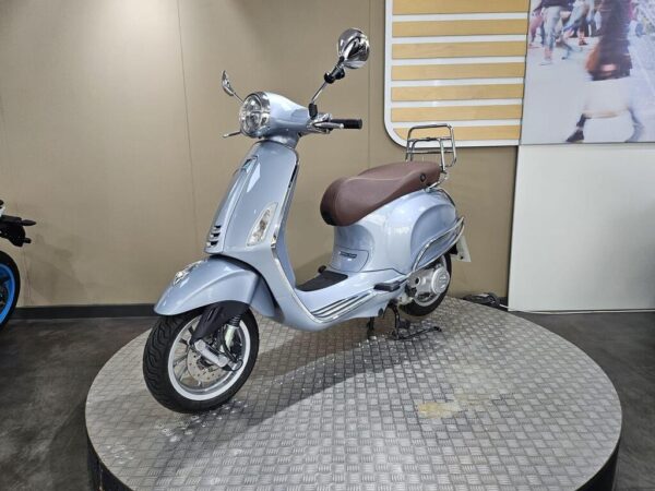 A blue scooter with a brown seat is displayed on a circular platform, featuring a shiny design and chrome details, set against a neutral background.