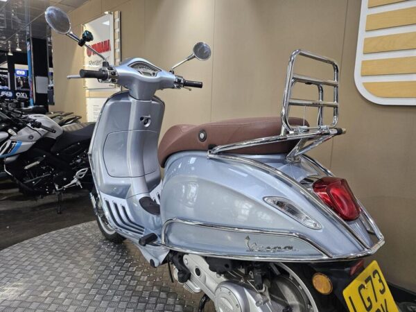 A silver Vespa scooter with a brown seat and chrome luggage rack, parked in a showroom with other motorcycles in the background.