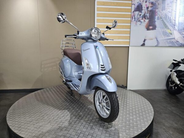 A light blue Vespa scooter is displayed on a rotating platform with a textured surface, featuring chrome mirrors and a brown seat, against a neutral background.
