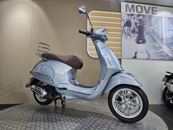 A light blue scooter with a brown seat is displayed on a round platform in a showroom, with a blurred city scene in the background and the word "MOVE" on the wall.