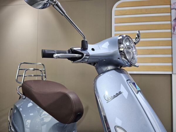 A close-up of a vintage-style scooter showcasing the handlebars, mirror, and headlight, with a brown seat and shiny chrome accents, set against a neutral background.