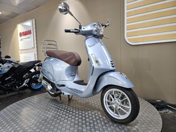 A light blue Vespa scooter with a brown seat is positioned on a diamond-patterned surface, with motorcycles in the background and a Yamaha logo on the wall.