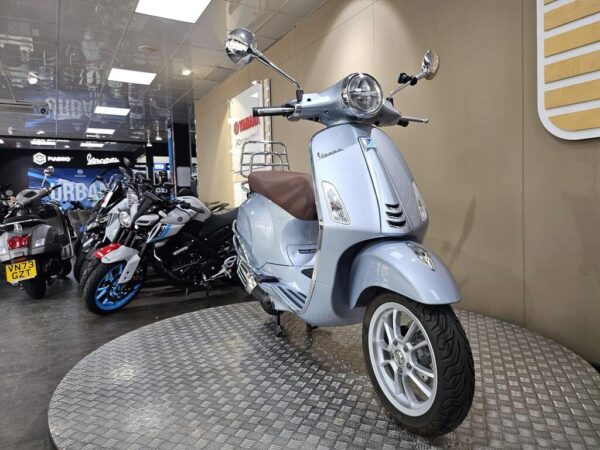A silver Vespa scooter displayed on a textured platform in a showroom, surrounded by various motorcycles in the background.