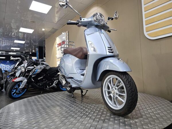 A blue Vespa scooter displayed on a metal floor in a motorcycle showroom, with several motorbikes visible in the background.