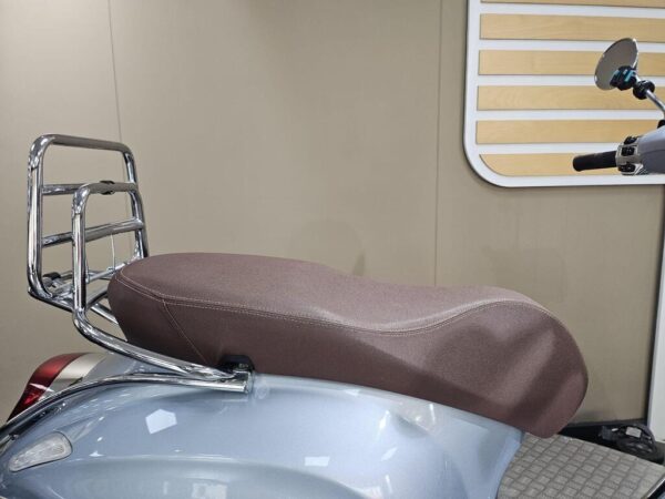 A close-up view of a motorcycle seat with a brown fabric covering and a chrome luggage rack attached to the back, set against a neutral-colored wall.