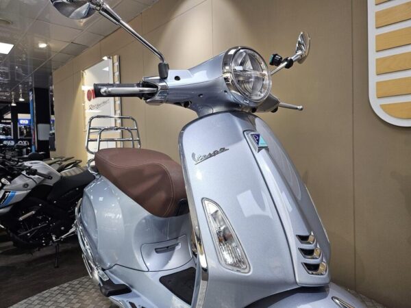 A close-up view of a silver Vespa scooter with a brown seat, showcasing its front design, handlebars, and headlight, set against a neutral background in a showroom.