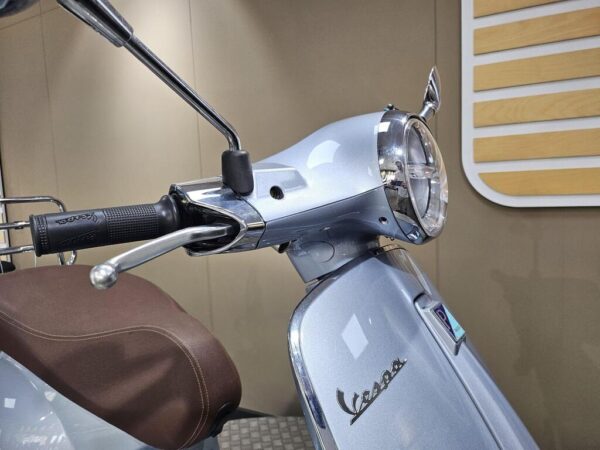 A close-up view of a silver Vespa scooter's handlebar, highlighting the chrome grips, headlight, and distinctive Vespa logo against a neutral background.