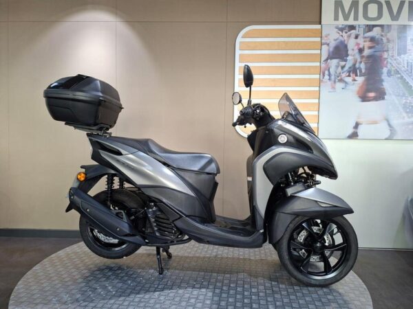 Yamaha Tricity (2017) A sleek gray scooter with a top case, displayed on a circular textured platform in a modern indoor setting, with a blurred background featuring people walking.