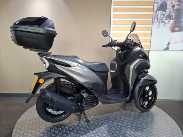 A sleek gray scooter with a rear top box, displayed on a circular metal platform against a neutral background.