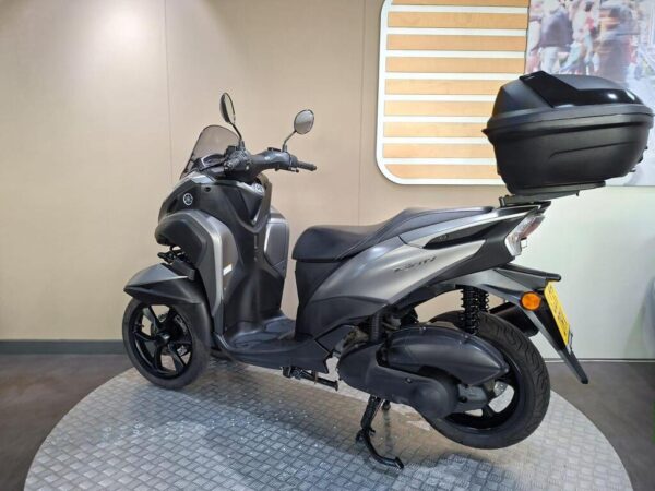 A gray scooter with a black top case parked on a circular platform in a showroom, featuring a windshield and dual mirrors. The background consists of neutral-colored walls.