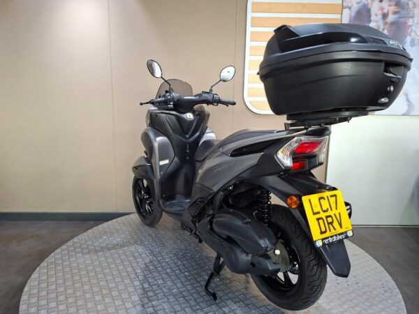 A black scooter featuring a top case, displayed on a circular platform with a plain background. The scooter is angled to show the rear and side view, highlighting its sleek design and modern features.