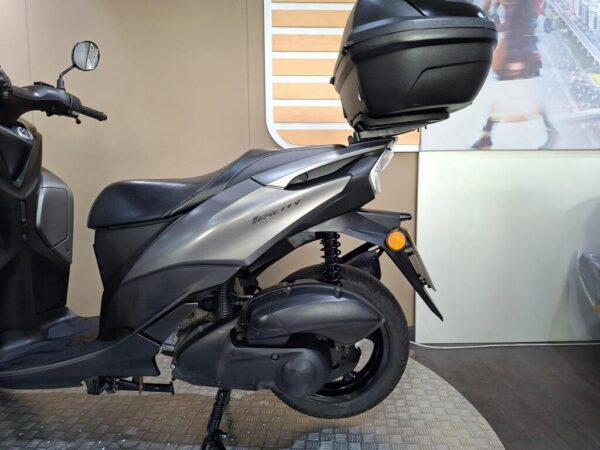 A side view of a gray scooter with a top box, parked on a textured floor, against a neutral background.