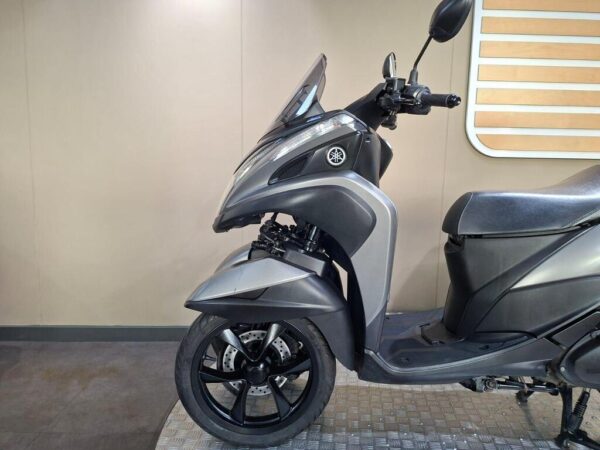 A side view of a grey Yamaha scooter parked on a textured surface, with a simple beige background and minimal decor.