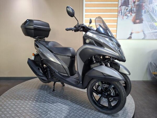 A gray three-wheeled scooter parked on a display stand in a showroom, featuring a rear storage box and modern design elements.