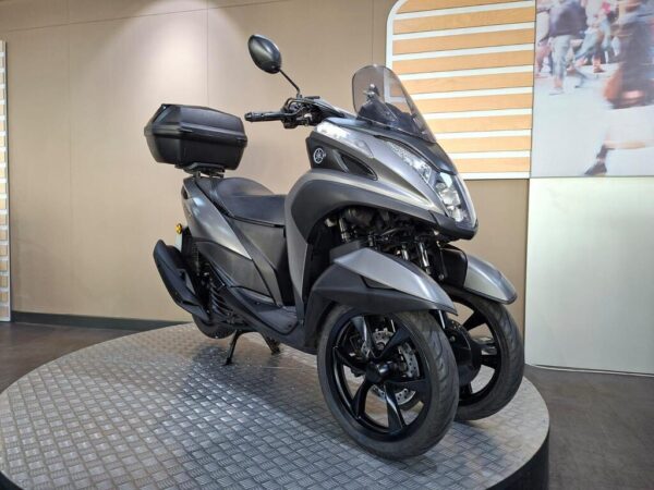 A modern grey scooter with three wheels, featuring a windscreen and a rear storage compartment, displayed on a round, textured platform against a neutral background.