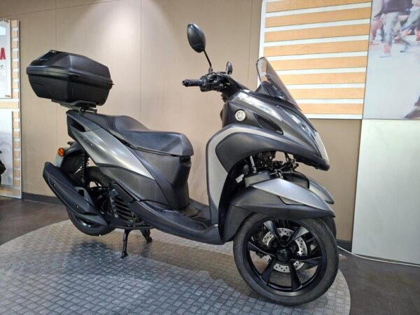A modern gray scooter with a black storage box on the back, displayed on a textured circular platform in a showroom.