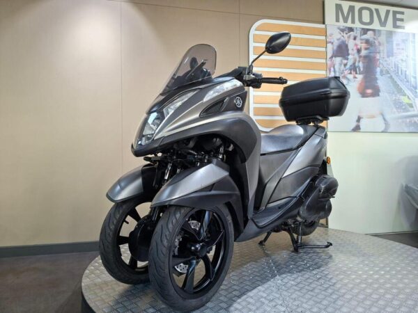 A sleek gray three-wheeled scooter displayed on a textured platform, set against a neutral wall featuring a "MOVE" sign and a lifestyle image in the background.
