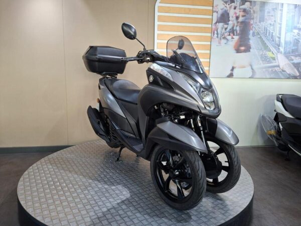 A gray three-wheeled scooter displayed on a circular platform in a showroom, with a modern design and a rear storage compartment. In the background, there is a blurred image of a busy city street.